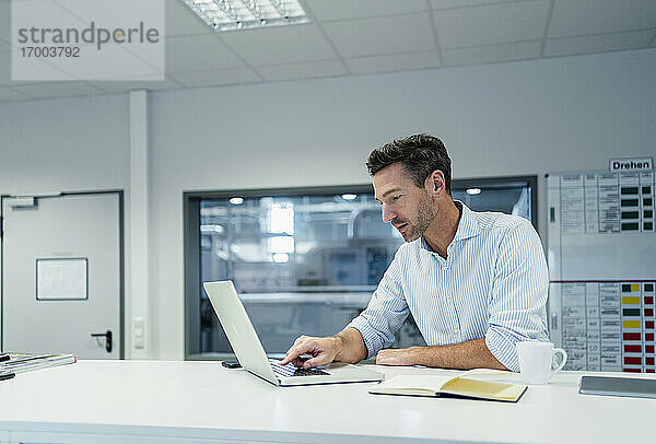 Männlicher Fachmann  der an einem Laptop arbeitet  während er im Sitzungssaal eines Fabrikbüros sitzt