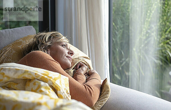 Reife Frau schaut durch das Fenster  während sie zu Hause auf dem Sofa liegt