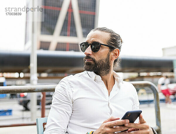 Porträt eines bärtigen Geschäftsmannes mit Sonnenbrille  der im Freien sitzt und ein Smartphone in der Hand hält