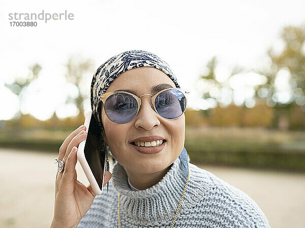 Junge Frau mit Kopftuch und Sonnenbrille lächelt  während sie im Freien mit einem Mobiltelefon spricht