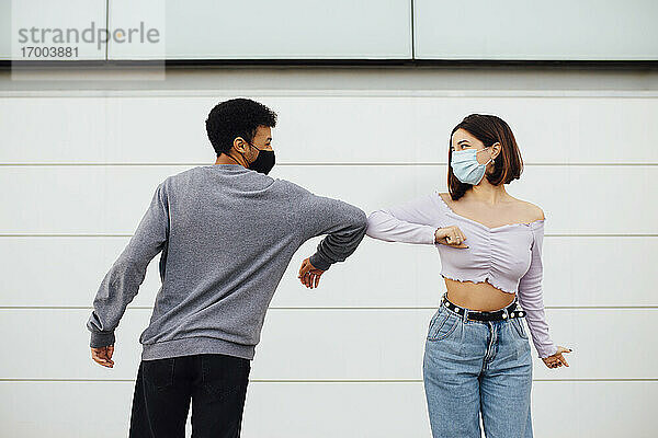 Junges Paar mit Gesichtsmaske grüßt mit Ellbogenstoß  während es an der Wand steht