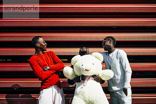 Drei lachende junge Männer mit riesigem Teddybär an roter Wand