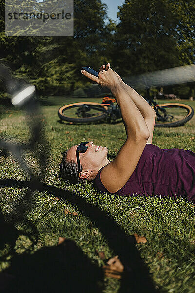 Sportlerin  die ein Mobiltelefon benutzt  während sie sich mit einem elektrischen Mountainbike im Park auf dem Rasen ausruht