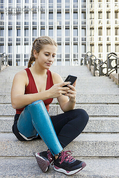 Sportlerin mit Beinprothese benutzt Mobiltelefon  während sie auf einer Treppe in der Stadt sitzt