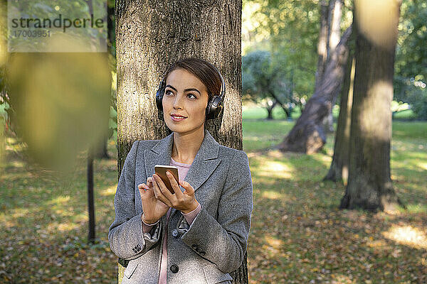 Nachdenkliche Geschäftsfrau hört Musik und hält ihr Smartphone im Herbst im Park