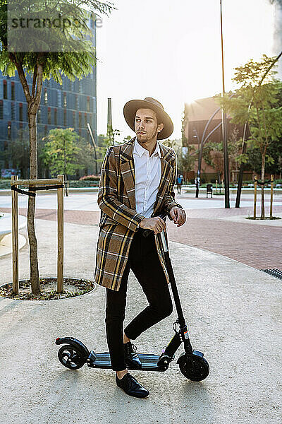 Stilvoller Geschäftsmann mit Hut und karierter Jacke steht auf einem Motorroller in der Stadt