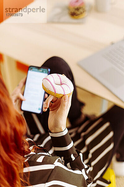 Geschäftsfrau  die einen Donut hält  während sie im Büro sitzt und ein Mobiltelefon benutzt