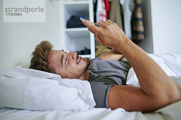 Junger Mann benutzt sein Smartphone  während er auf dem Bett im Schlafzimmer liegt