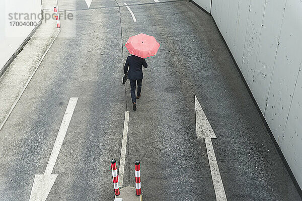 Rückansicht eines Geschäftsmannes mit Regenschirm  der auf einer Straße mit Pfeilschildern geht