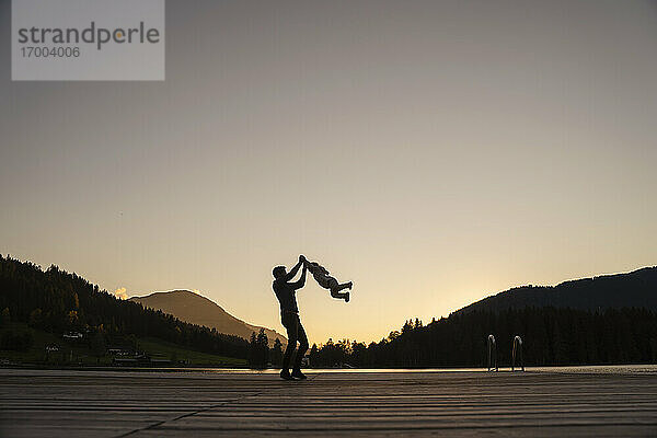 Silhouetten von Vater und kleiner Tochter  die in der Abenddämmerung am Ende eines Stegs am Seeufer spielen