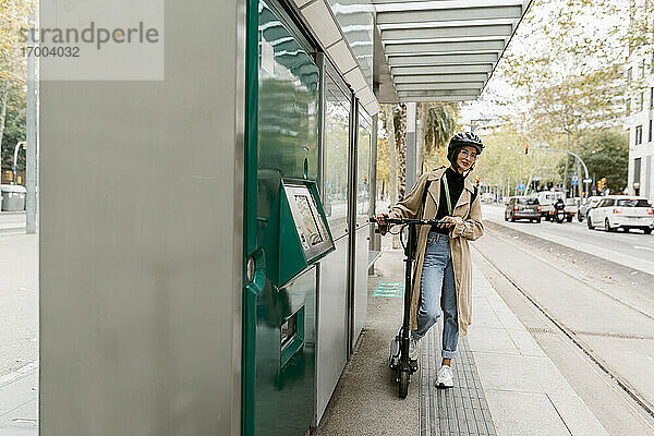 Frau mit Fahrradhelm  die mit einem Elektroroller an einer Straßenbahnhaltestelle in der Stadt wartet