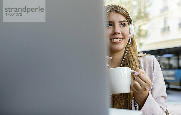 Lächelnde junge Frau mit Laptop und Kaffeetasse  die wegschaut  während sie in einem Straßencafé sitzt