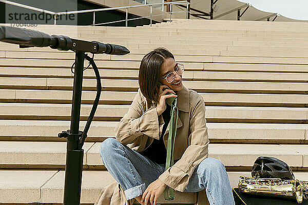 Lächelnde Frau  die mit einem Mobiltelefon spricht  während sie neben einem Saxophoninstrument und einem elektrischen Roller auf einer Treppe sitzt