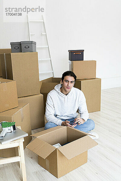 Junger Mann mit Mobiltelefon sitzt bei Kartons in neuer Wohnung