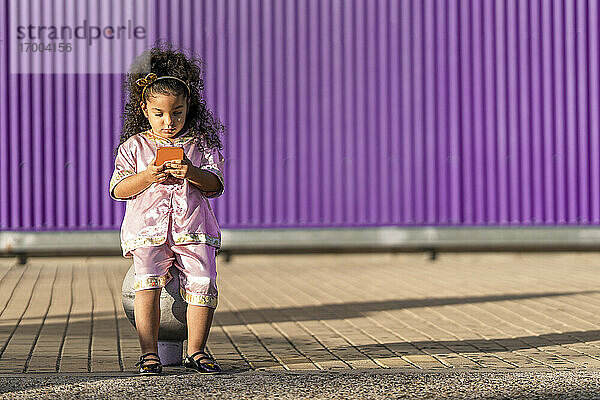 Nettes Mädchen  das ein Mobiltelefon benutzt  während es an einer lila Wand sitzt