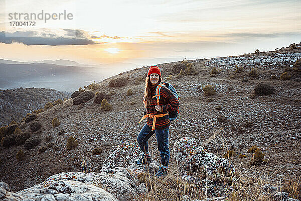 Lächelnde Wanderin  die wegschaut  während sie bei Sonnenuntergang auf einem Felsen steht