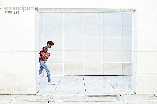 Junge Frau hält roten Ballon in Herzform  während sie gegen eine weiße Wand läuft