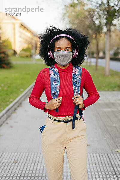 Frau mit Schutzmaske und Kopfhörern  die einen Rucksack trägt  während sie auf einem Fußweg steht