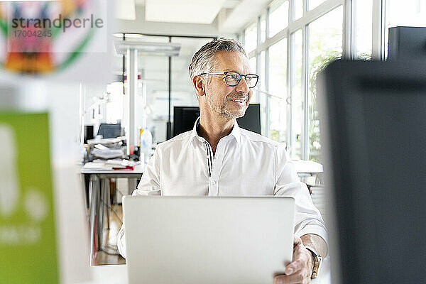 Lächelnder männlicher Fachmann  der mit einem Laptop im Büro sitzt und wegschaut