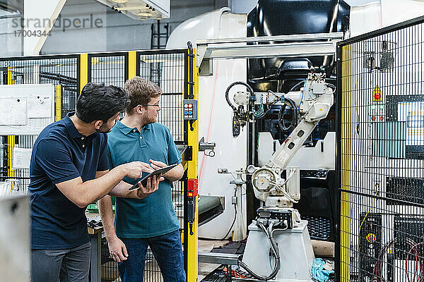 Männliche Mitarbeiter  die ein digitales Tablet halten und einen Roboterarm betrachten  während sie in der Fertigungsindustrie stehen