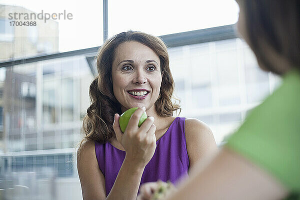 Lächelnde Geschäftsfrau  die einen Apfel in der Hand hält und eine Kollegin in der Büro-Cafeteria ansieht