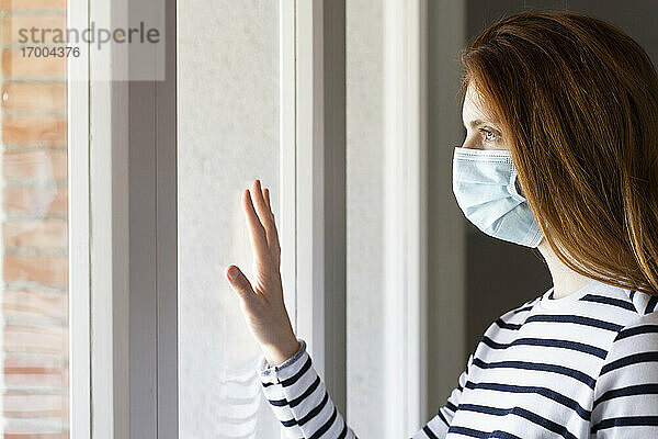 Rothaarige Frau mit Gesichtsschutzmaske steht während COVID-19 am Fenster ihres Hauses
