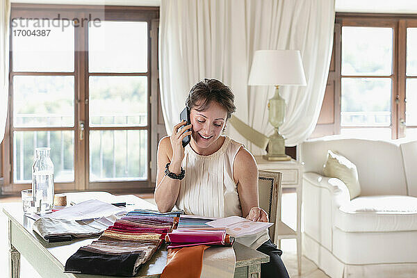 Geschäftsfrau  die mit ihrem Smartphone telefoniert  während sie zu Hause Farben aus Stoffmustern auswählt