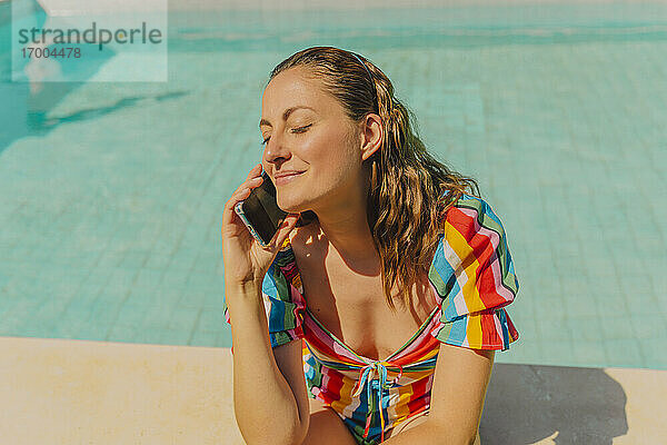 Porträt einer glücklichen Frau am Telefon  die am Pool sitzt