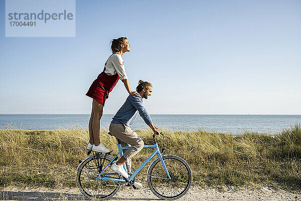 Freundin genießt Fahrt mit Mann auf dem Fahrrad stehend gegen klaren Himmel