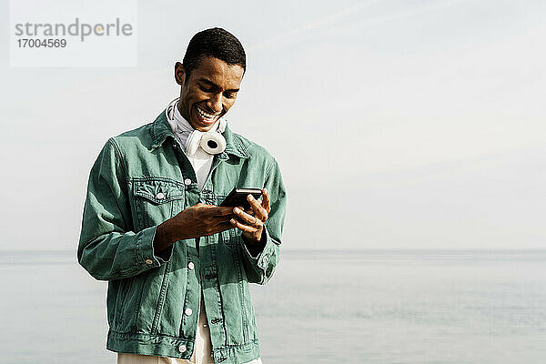 Lächelnder junger Mann mit Smartphone gegen den Himmel