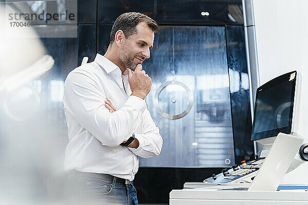 Lächelnder männlicher Unternehmer mit Hand am Kinn  der auf einen Laptop schaut  während er in einer Fabrik steht