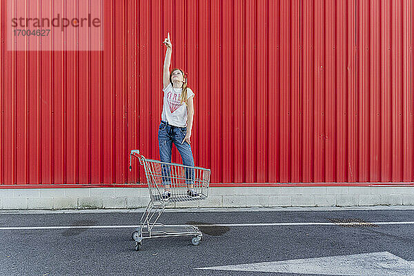 Mädchen in einem Einkaufswagen zeigt mit dem Finger nach oben vor einer roten Wand