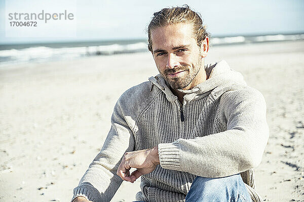 Lächelnder Mann mit Hand auf dem Knie am Strand sitzend