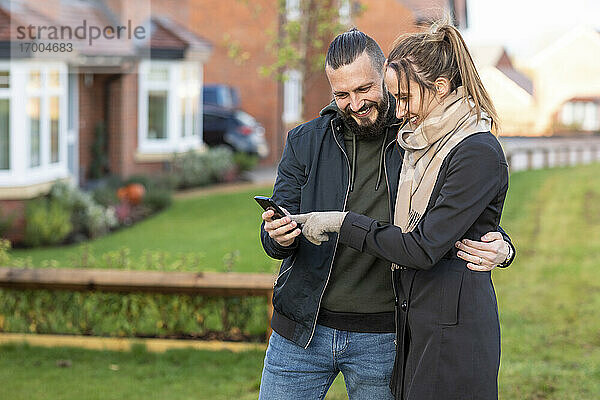 Glückliches Paar schaut auf sein Smartphone  während es im Vorgarten steht
