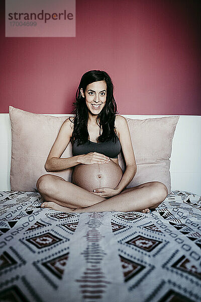 Lächelnde schwangere Frau sitzt auf dem Bett gegen die rote Wand zu Hause