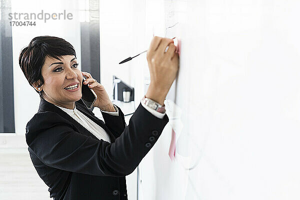 Porträt einer Geschäftsfrau  die mit einem Smartphone spricht und auf einem Klebezettel schreibt  der an einer Tafel im Büro hängt