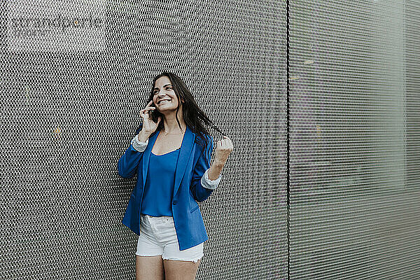 Glückliche Unternehmerin  die mit einem Smartphone spricht  während sie an der Wand steht
