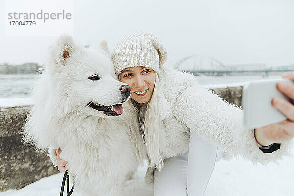 Weiß gekleidete Frau nimmt Selfie mit ihrem weißen Hund im Winter