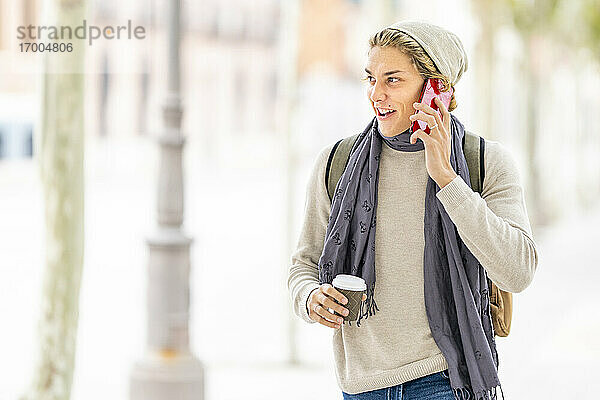 Lächelnder Mann mit Kaffeetasse  der beim Spazierengehen in der Stadt mit seinem Mobiltelefon spricht