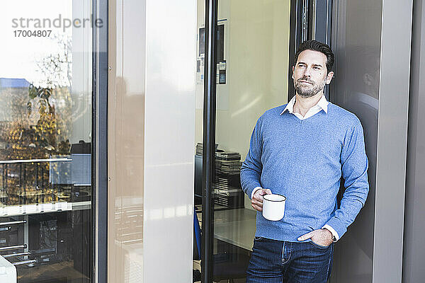 Geschäftsmann hält eine Kaffeetasse und lehnt sich mit der Hand in der Tasche am Eingang eines Büros an die Wand