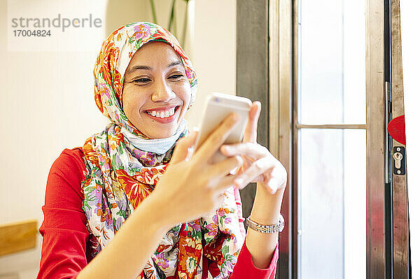 Glückliche malaysische Frau mit Hidschab  die in einem Café ein Smartphone benutzt