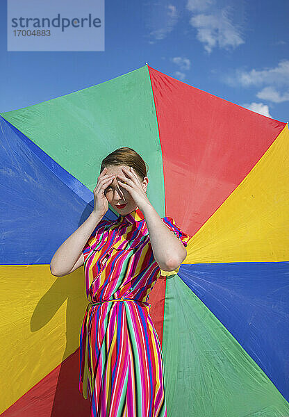 Frau  die ihre Augen abschirmt  während sie vor einem bunten Sonnenschirm steht