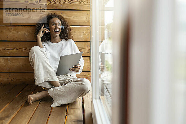 Lächelnde Frau mit Kopfhörern und digitalem Tablet  während sie zu Hause am Fenster sitzt