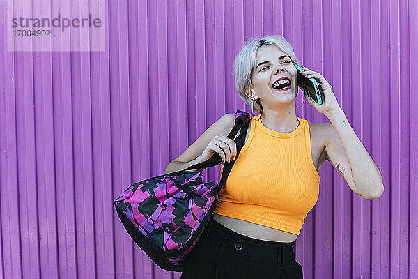 Blonde Frau mit Tasche  die ihr Smartphone vor einer lila Wand benutzt