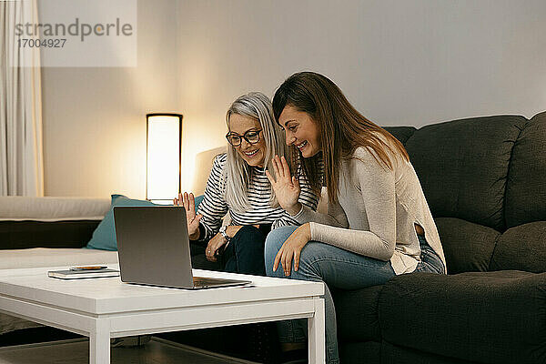 Lächelnde Frauen winken mit der Hand zum Videoanruf auf dem Laptop  während sie zu Hause auf dem Sofa sitzen