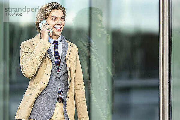 Junger Geschäftsmann im Anzug  der sich an eine Glaswand lehnt und mit einem Mobiltelefon spricht