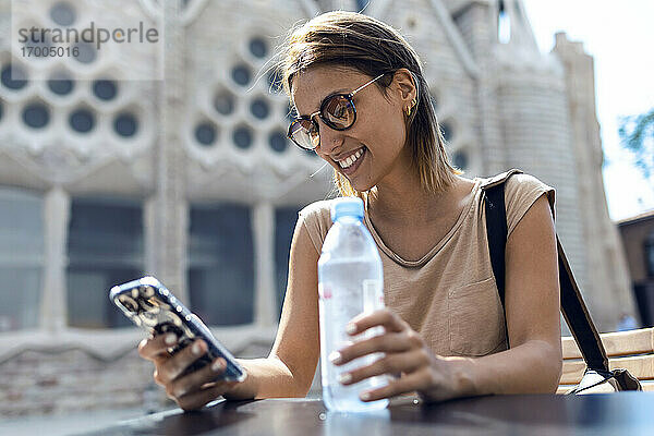 Junge Frau mit Sonnenbrille  die ein Mobiltelefon benutzt  während sie vor der Sagrada Familia in Barcelona  Katalonien  Spanien sitzt