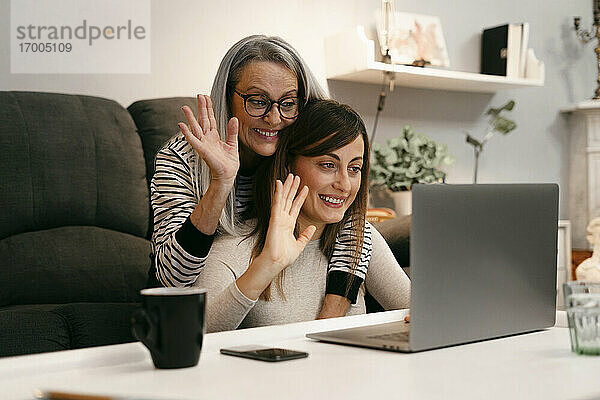Glückliche Frauen winken mit der Hand zum Videoanruf auf dem Laptop  während sie zu Hause sitzen