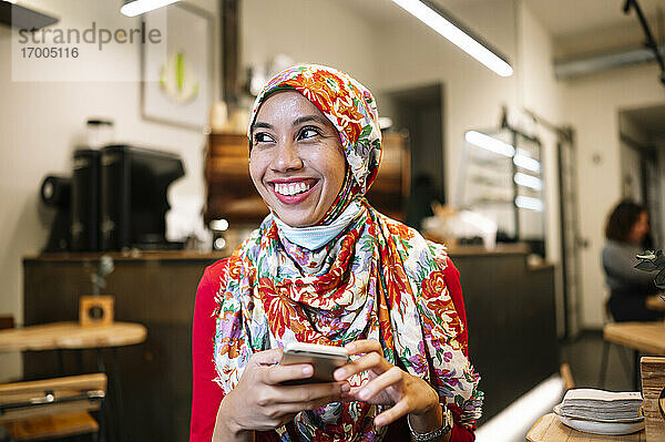 Lächelnde muslimische Frau in geblümtem Hidschab  die wegschaut  während sie in einem Café ein Mobiltelefon hält