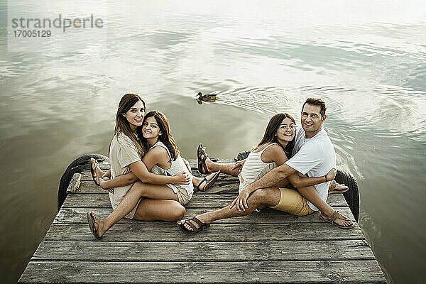 Töchter umarmen Mutter und Vater  während sie auf einem Steg am See sitzen